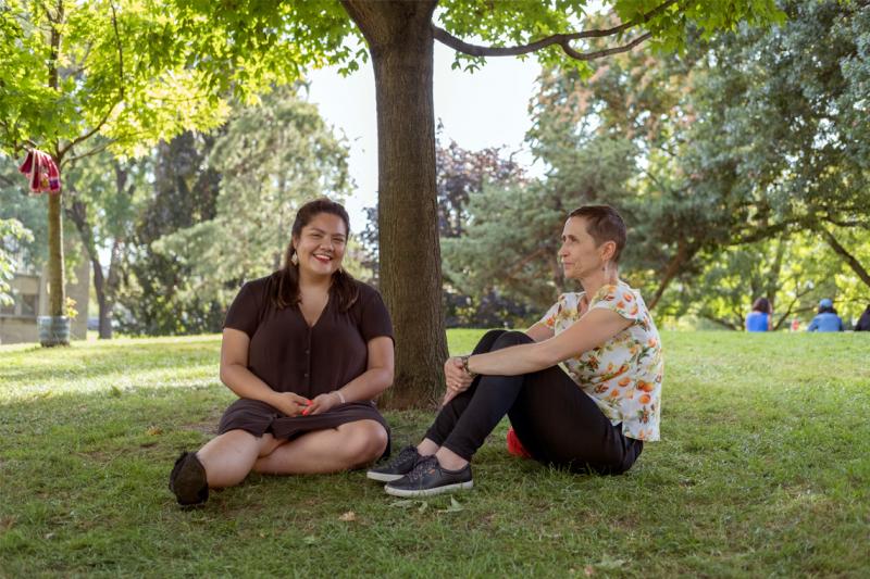 Kateri Lucier-Laboucan and Andrea Mantin smile together, sitting on a lawn under a tree.