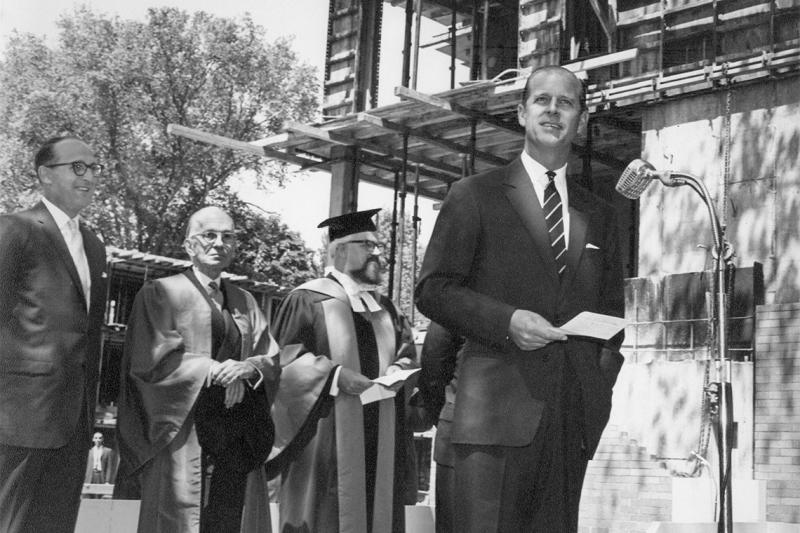 In a photo from 1962, a young Prince Philip speaks at a microphone on a building site, with men in academic robes behind him.