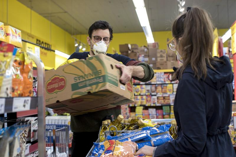In a grocery store, Adam Zivo lifts a cardboard box full of groceries. He is wearing a mask.