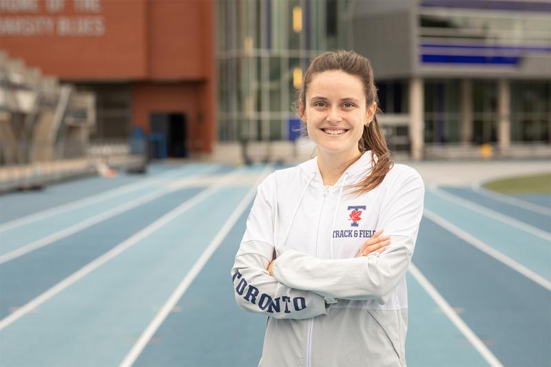 Lucia Stafford smiles as she stands with arms crossed on the Varsity Stadium track. She wears a Varsity Blues hoodie.