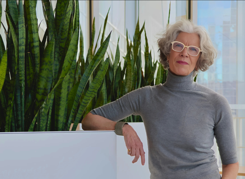 A woman in a grey turtleneck and glasses poses next to some potted plants