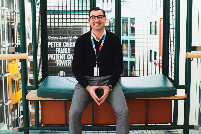 Michael Gritti smiles, as he sits in a balcony alcove with a large atrium behind him.