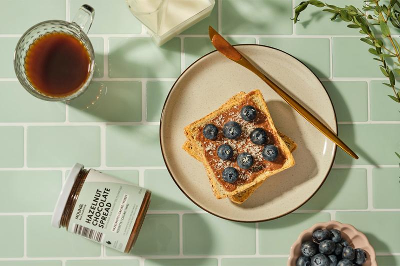 A slice of toast with chocolate spread and blueberries, beside a jar labelled Mounib Hazelnut Chocolate Spread.