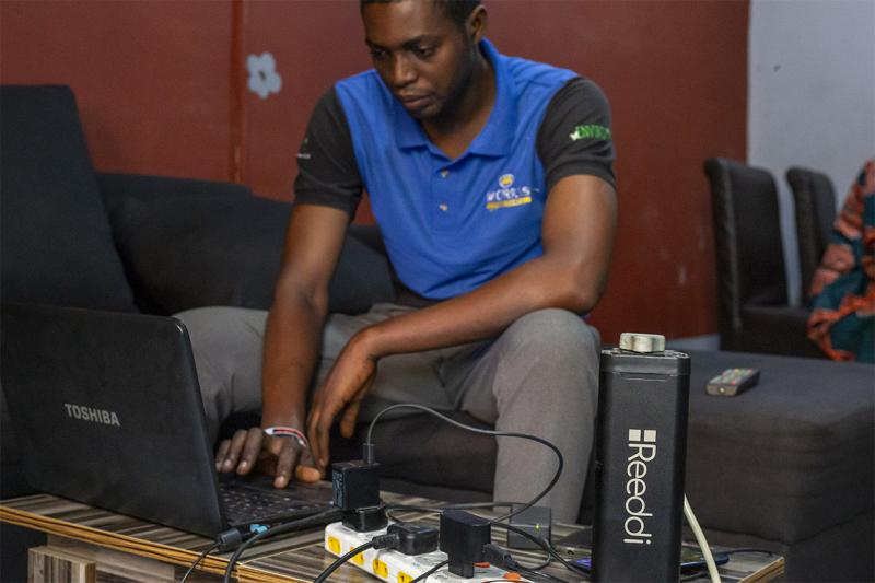 A young man types on a laptop that is plugged into a milk-carton-sized battery pack. The name Reeddi is written on the pack.