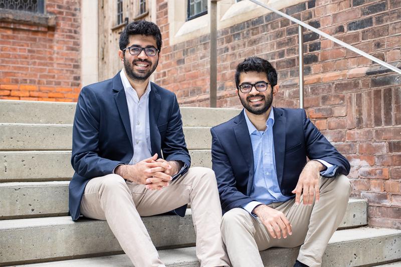 Nishul and Nakul Juneja, dressed identically, laugh as they sit on steps outside a building.