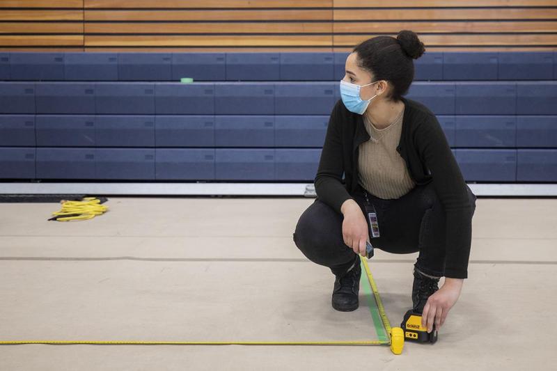 Reem El-Ajou, wearing a mask, uses a tape measure to mark off a social distancing line on the floor of a gymnasium.