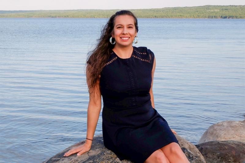 Shanna Peltier smiles, sitting on a large rock on the shores of a lake.