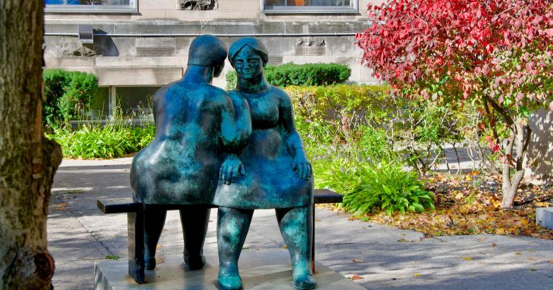 In the garden at St. Michael's College, a metal sculpture depicts two women on a bench chatting together.