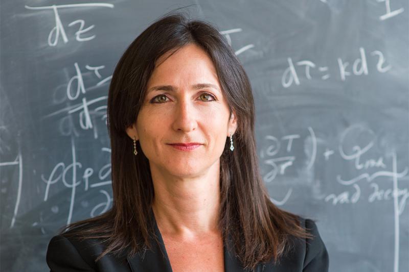 Sara Seager smiles quietly, standing in front of a blackboard covered with equations.