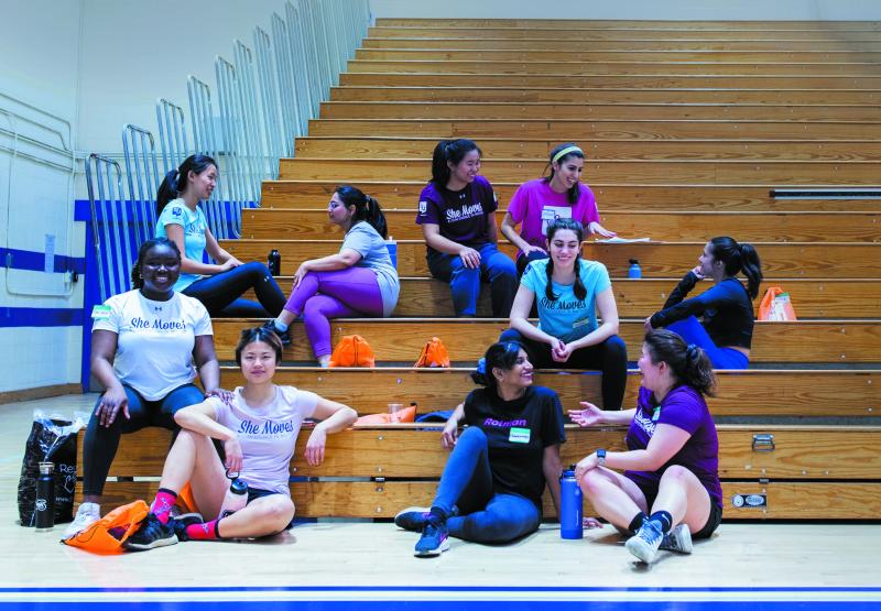 A diverse group of smiling students chat on the bleachers in a gym, wearing She Moves t-shirts.