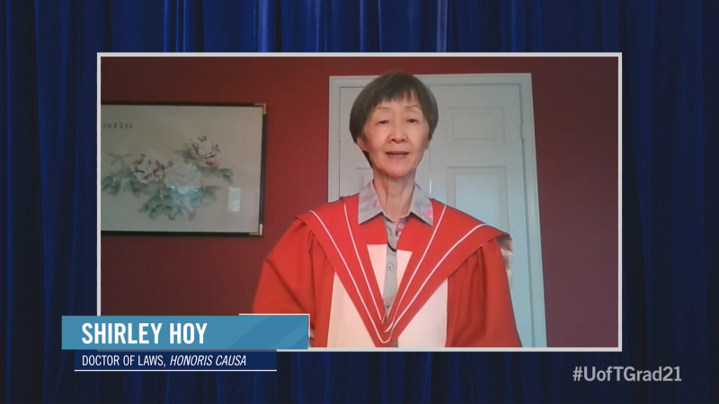 Shirley Hoy, wearing academic robes, speaks on video while standing in front of a painting of flowers.