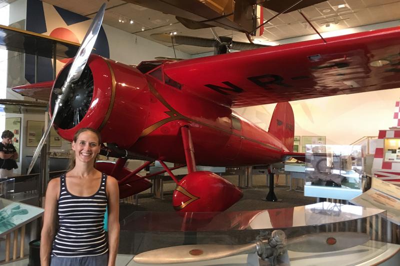 Lindsay Zier-Vogel stands by a small airplane with a single nose-propellor and teardrop-shaped wheel covers.