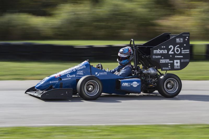 The half-sized U of T Formula SAE racing car speeds down a track. It features an open cockpit, rollbar, and huge rear wing.