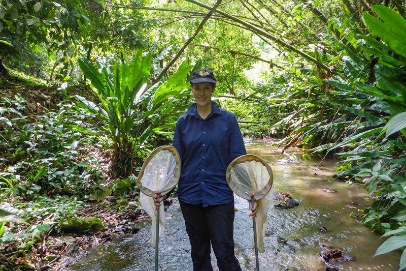 Alex De Serrano stands in a shallow stream, holding two scoop-shaped nets. Lush vegetation arches overhead.