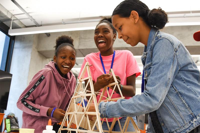 Three young girls laugh with delight as they construct a model bridge out of popsicle sticks.
