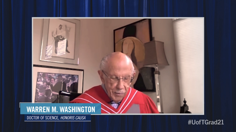 Warren Washington, wearing academic robes, speaks on a video call from a room decorated with paintings.
