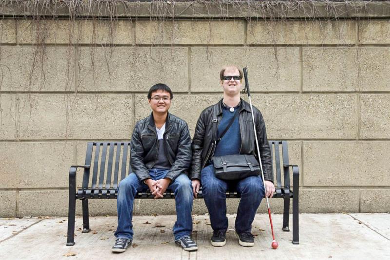 Rylan Vroom, who uses a white cane, and Bin Liu, who does not, smile as they sit together on a bench.