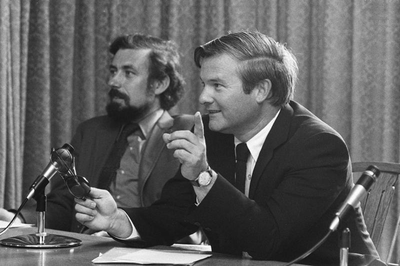 In a photograph from the 1970s, Bill Davis leans on a table and speaks into a microphone.