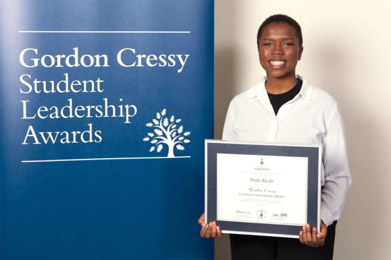 Dede Akolo smiles and holds a certificate next to a sign reading: Gordon Cressy Student Leadership Awards.