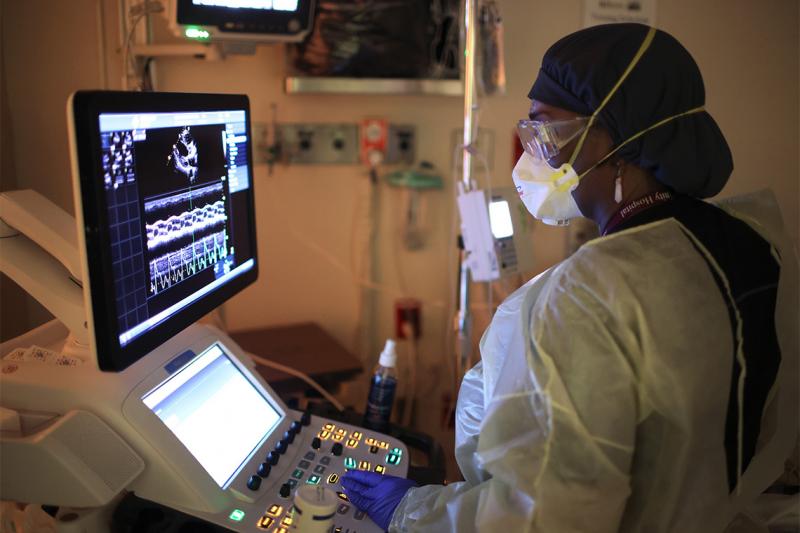 A masked health worker reads a screen. The screen shows a streaky image of a heart, surrounded by graphics and numbers.