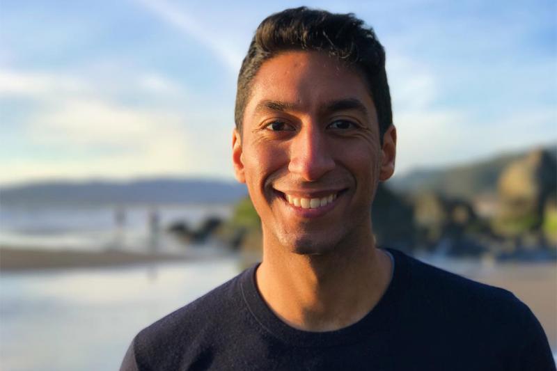 Zain Manji smiles, standing in front of a wooded lake at sunset.