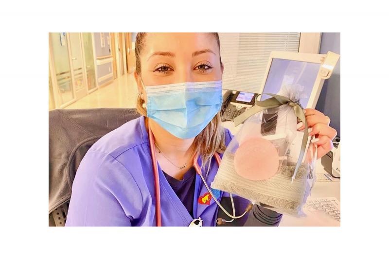 A young woman wearing a mask and scrubs holds up a bag containing a towel and a tube of a skin-care cream.