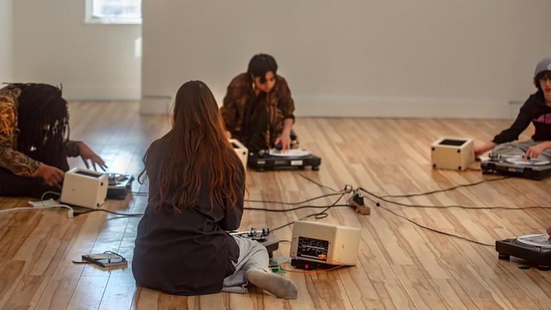 A group of young people sitting on a hardwood floor with some music equipment