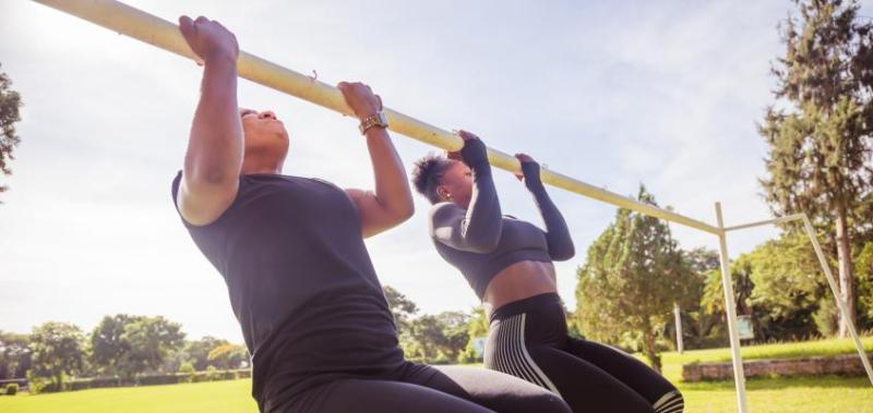 Two women in a park in Zambia, wearing athletic gear, do pull-ups on a bar.