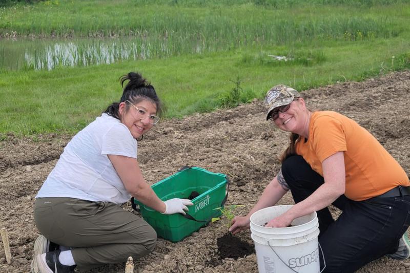 Lisa Owl and Melanie Jeffrey smile as they kneel and place a crookneck squash seedling in the earth.