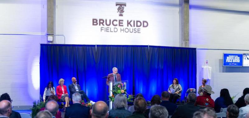 An audience gathers to hear Bruce Kidd speak, beneath a sign that reads, Bruce Kidd Field House.