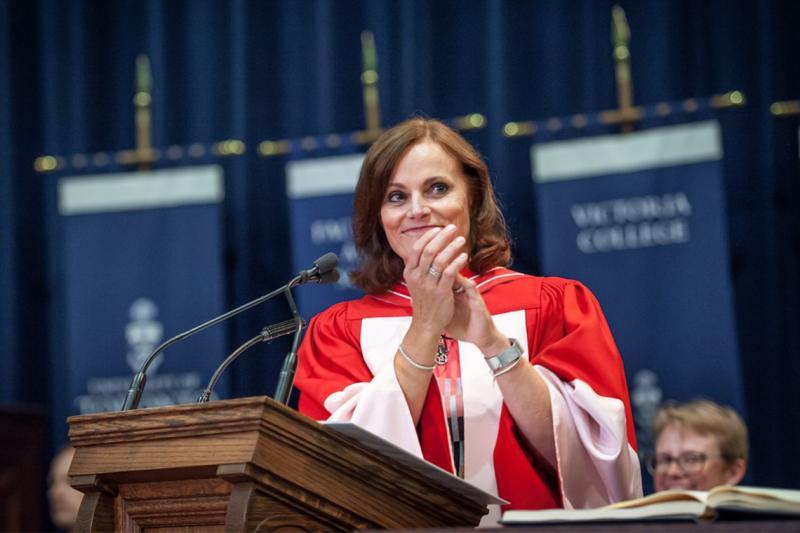 Judy Goldring smiles and claps as she stands at a podium, wearing academic robes.