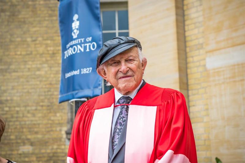 Nate Leipziger, wearing academic robes and his trademark leather cap, stands outside Convocation Hall.