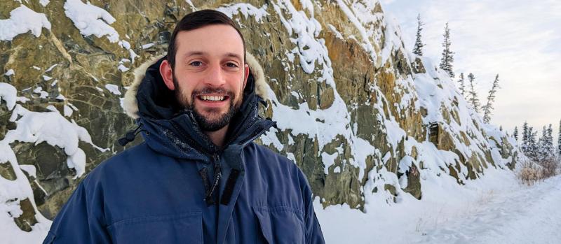 Luke Spooner smiles, as he stands in front of a snow-covered cliff, in a spruce forest.