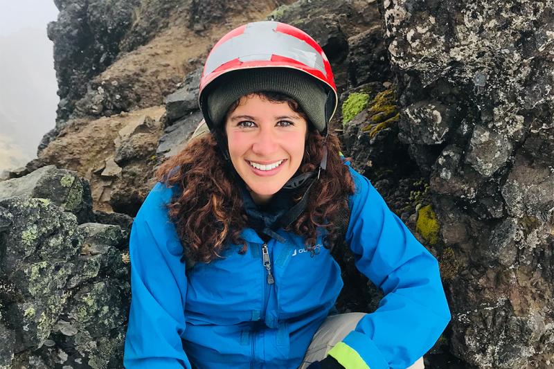 Marah Ayad smiles. She is wearing a hard hat and hiking gear and standing in front of a rocky cliff face.