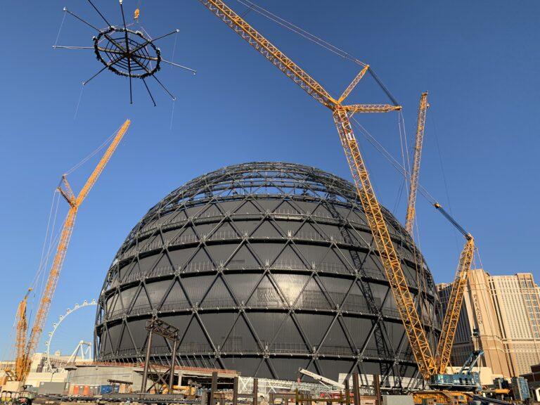 A crane lifts a spiderweb-shaped object high above a huge sphere built out of triangles.