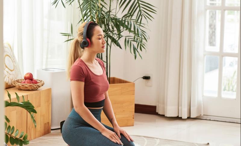 A woman wearing headphones kneels on a yoga mat in a living room.