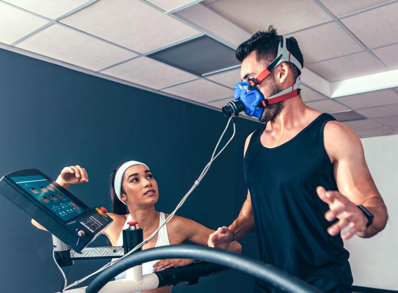 A young man runs on a treadmill, wearing a mask to measure his breathing. A young woman operates the controls.