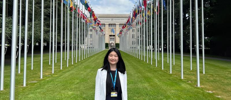 Yifan Zhou smiling. standing next to the rows of flags outside the United Nations building in Geneva.