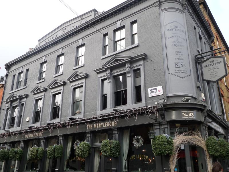 An exterior photo of the Marylebone Pub, London. It's a grey building on the corner of Marylebone High street and Moxon Street. The former name of the pub is visible at the top, etched in the stone (The Queen's Head)