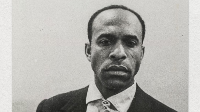 Portrait photography of Frantz Fanon in a suit and tie looking at the camera against a grey background.