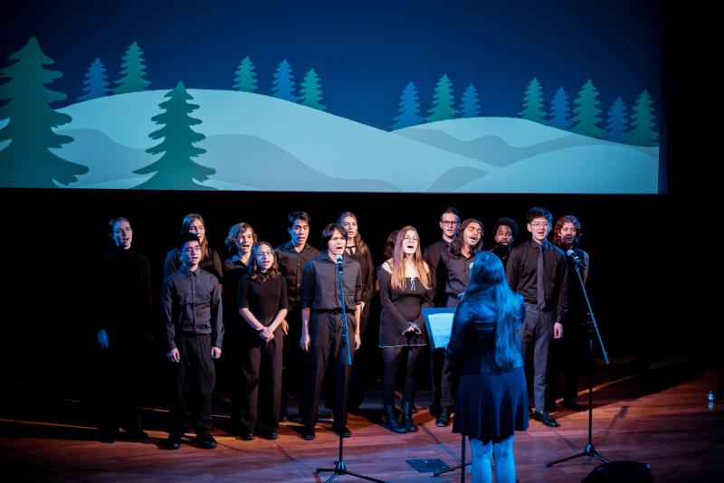 People dressed in black outfits, standing in front of an image of a mountain with pine trees, singing.