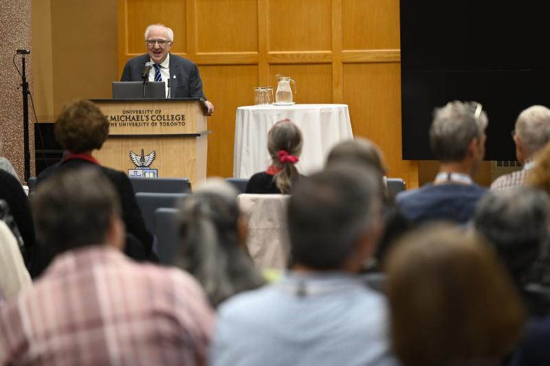 Douglas Moggach in a suit and tie speaks at a podium labeled "University of St. Michael’s College, The University of Toronto" in front of an audience. He is smiling while delivering lecture. The setting appears to be an academic or alumni event, with a table holding a water pitcher and glasses beside him and attendees seated and facing the speaker.