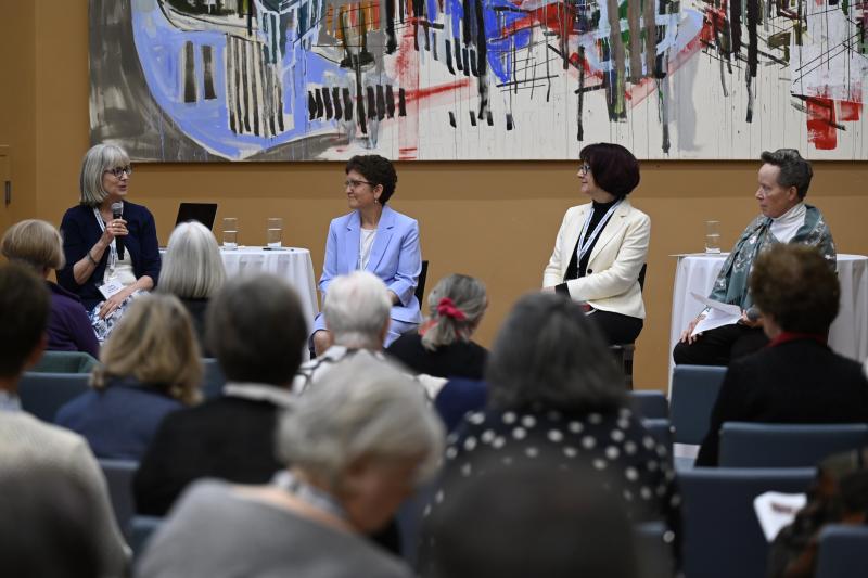"Four women sit at the front of a room engaged in a panel discussion at St. Michael’s College, University of Toronto. One of the women speaks into a microphone while the others listen attentively. They are seated in front of a colorful abstract painting, with round tables holding water glasses beside them. An audience of older adults listens from rows of chairs.