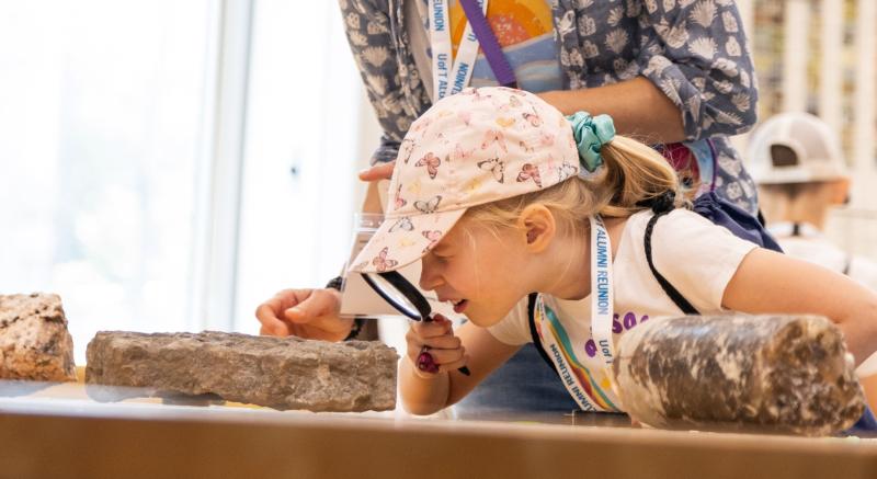 A young child, accompanied by an adult, examines a rock through a magnifying glass.