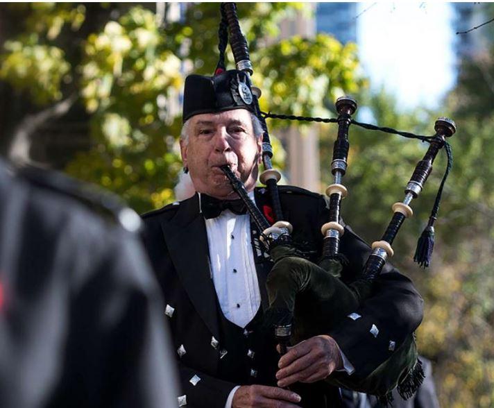 Man playing bagpipe, with trees behind him. Man dressed in black with white shirt. 