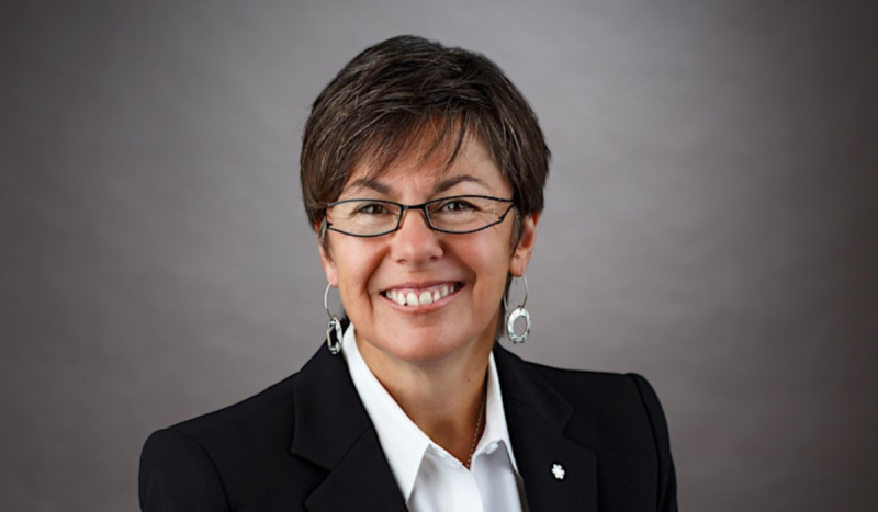 Photo of speaker Kim Pate wearing black jacket, white shirt, glasses against a grey background. 