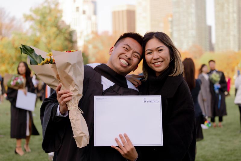 Graduate with parent, both dressed in black holding flowers and a certificate.