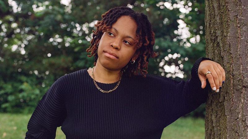 Isabel Okoro standing outside leaning their arm on a tree, while looking at the view of nature.
