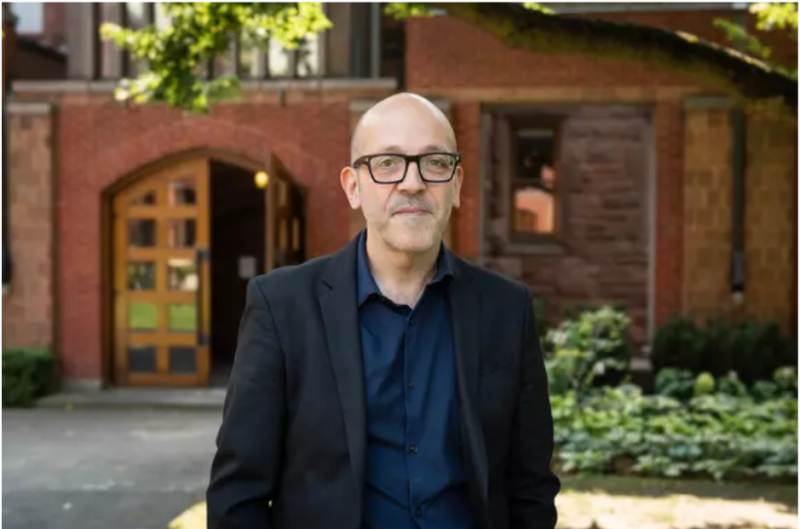 Image of man in shirt, suit and black rimmed eye glasses, standing outside with trees and building in background. 