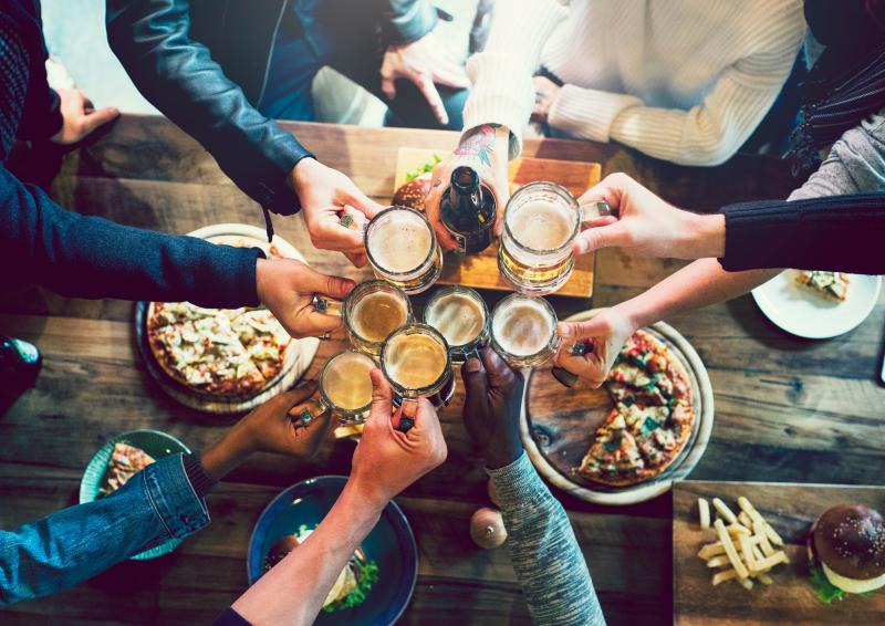 A group of friend cheersing glasses over a wood bar table full of snacks.